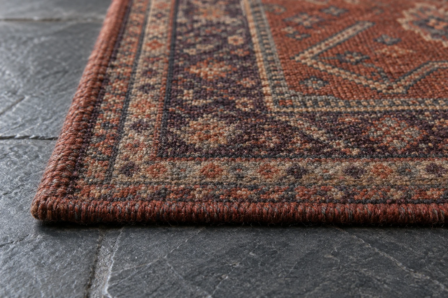 Close-up of a terracotta patterned wool rug pile and bound edge on a gray stone floor