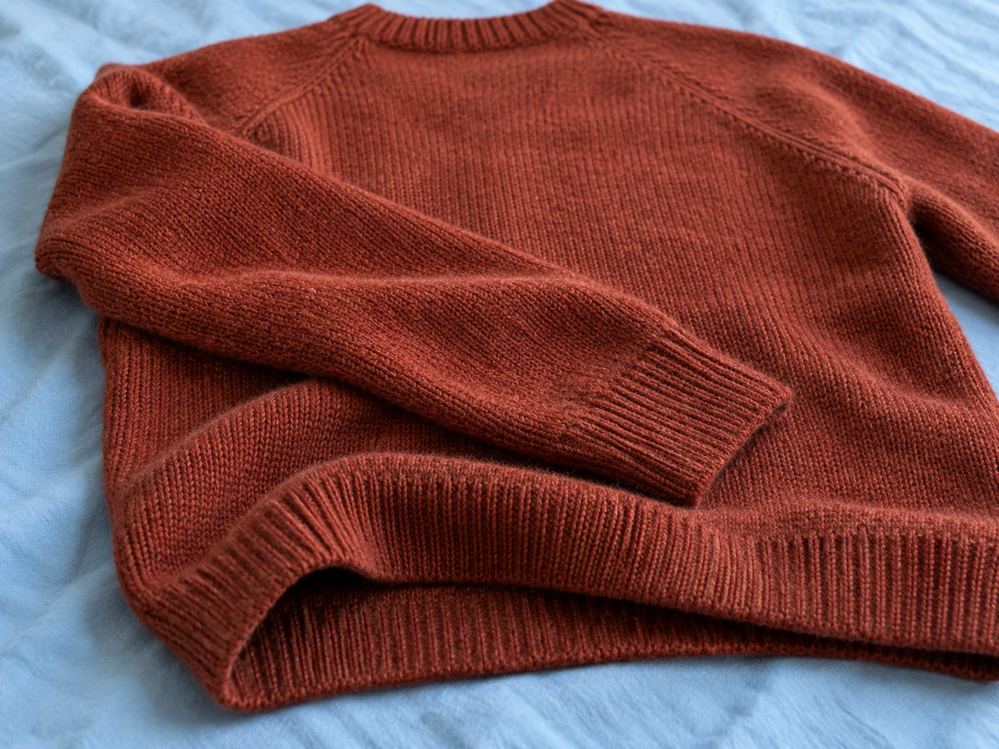 A rust-colored wool sweater resting in a loose fold on a pale blue cotton surface.